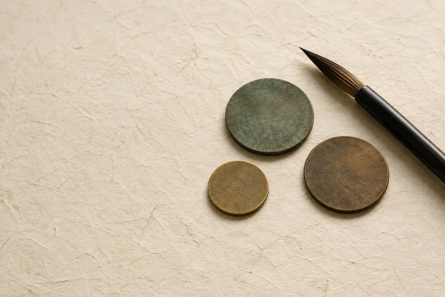 Three unmarked metal coins on textured paper beside an ink brush